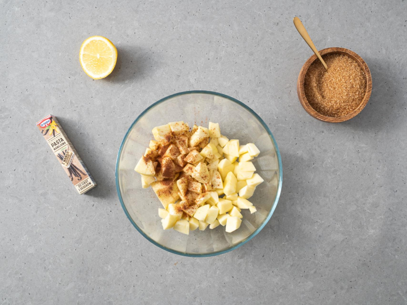 W miseczce obrane jabłka posypane cynamonem. Obok w opakowaniu pasta z wanilii , w miseczce cukier trzcinowy, a na blacie połówka soku z cytryny.