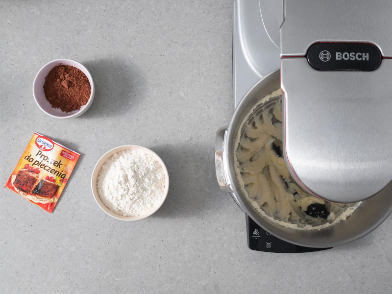 Ucieranie w misie robota kuchennego masła z cukrem. Obok proszek do pieczenia dr. oetkera, kakao oraz mąka pszenna.