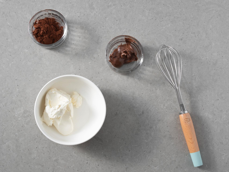 W miskach składniki do kremu czekoladowego na babeczki: serek śmietankowy, kakao i krem czekoladowy. Obok rózga kuchenna.