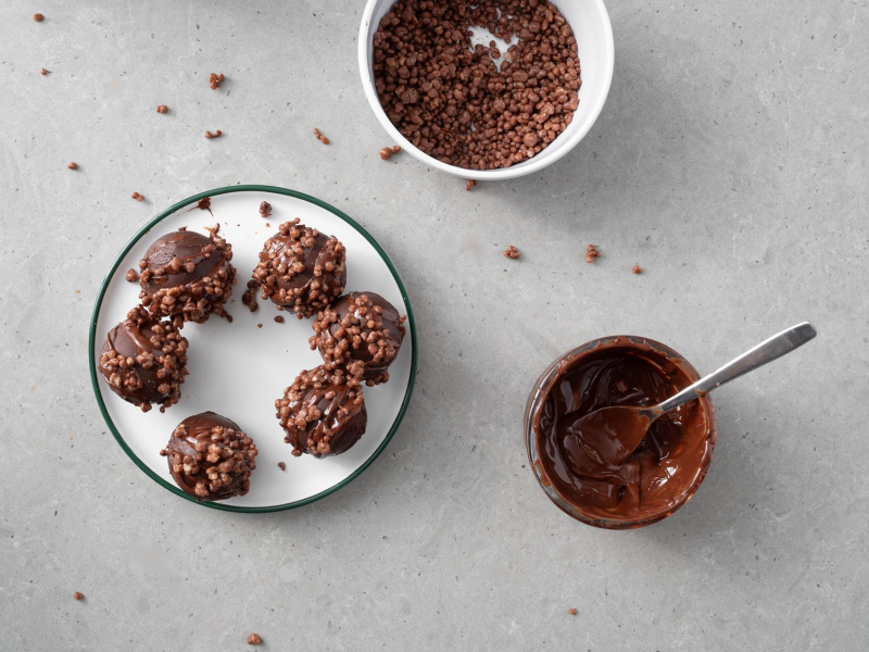 Gotowe kule z czekolady do mleka. Obok rozpuszczona w miseczce czekolada oraz dekoracyjne chrupki czekoladowe.