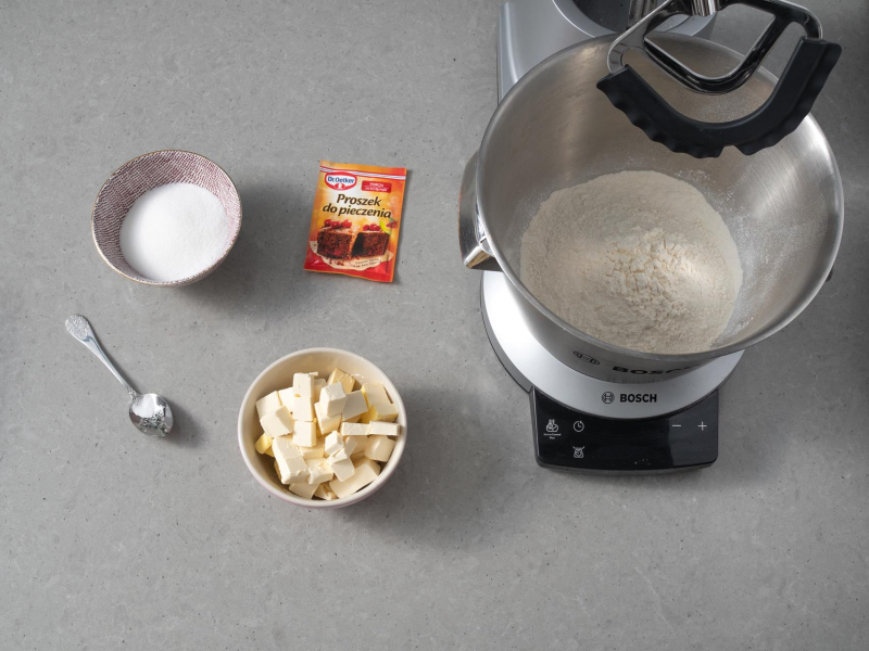 W misie misera firmy Bosch przesypana mąka. Obok w miseczce cukier, posiekana margaryna, proszek do pieczenia Dr. Oetker i sól na łyżeczce.