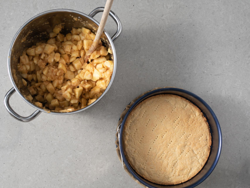 W garnku podprażone jabłka. Obok tortownica z upieczonym spodem do szarlotki.