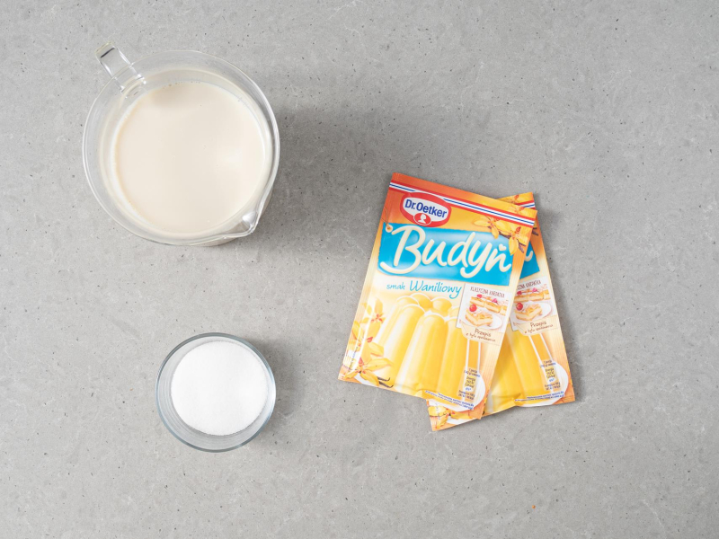 Na blacie dwa opakowania budyniu o smaku waniliowym dr. oetkera. Obok w miseczce cukier oraz napój sojowy.