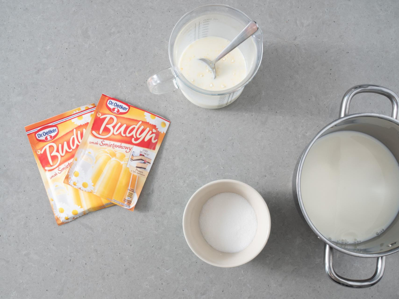 Na blacie kuchennym 2 opakowania budyniu o smaku śmietankowym. Obok garnek z mlekiem i miseczka z cukrem.