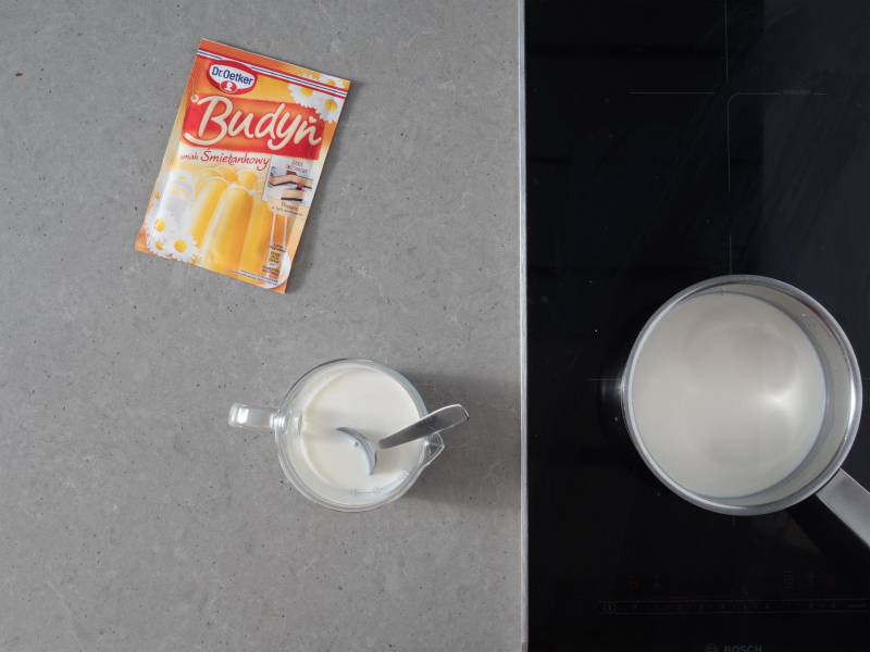 Na blacie budyń o smaku śmietanowym. Obok dzbanek z mlekiem i garnek na płycie indukcyjnej.