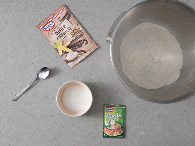W misie mąka pszenna. Obok na blacie opakowanie cukru z wanilią i drożdży. W miseczce cukier.