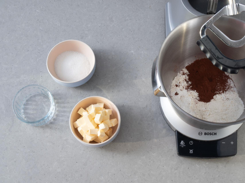Na blacie robot kuchenny firmy Bosch. Obok w miseczce cukier, masło i woda..