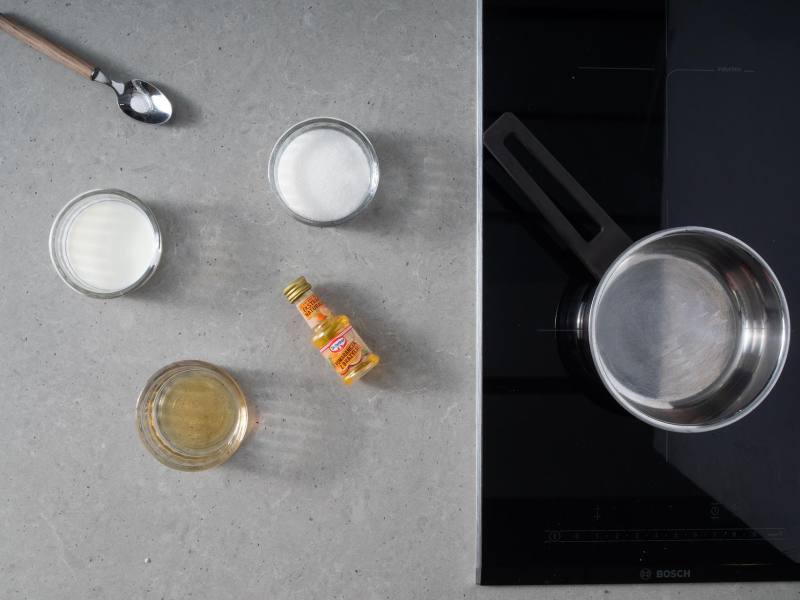 Na płycie indukcyjnej garnek. Obok na blacie kuchennym sól na łyżeczce, cukier, miód, mleko i ekstrakt pomarańczowy.