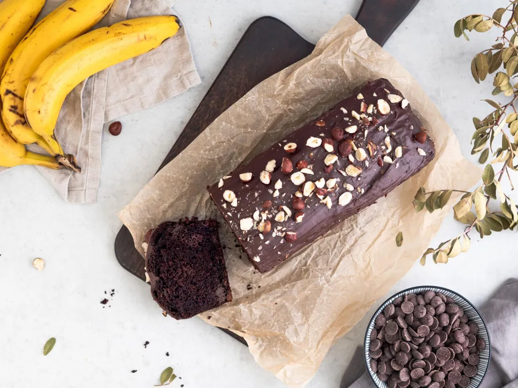Ciasto czekoladowe z bananami polane czekoladą i obsypane orzechami laskowymi na drewnianej desce - Przepis Wszystkiego Słodkiego