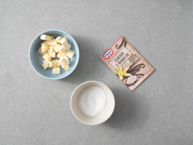 W miseczkach: pokrojone w kostkę masło roślinne i cukier. Obok opakowanie cukru z wanilią.