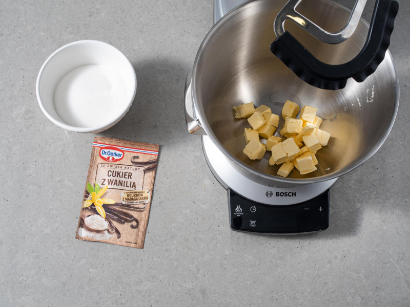 Margaryna roślinna pokrojona w kostkę w misie robota kuchennego firmy Bosch. Obok cukier w miseczce i opakowanie cukru z wanilią Dr. Oetkera.