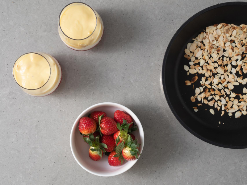 Truskawki w miseczce. Obok podpra偶one p艂atki migda艂贸w na patelni i pucharki z sosem waniliowym.