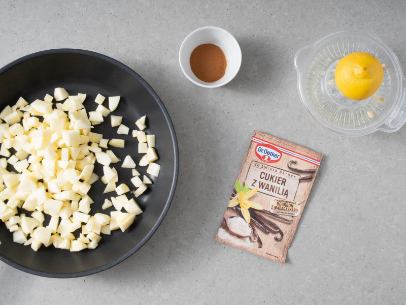 Na patelni pokrojone jabłka. Obok w miseczce cynamon, opakowanie cukru z wanilią oraz połówka cytryny.
