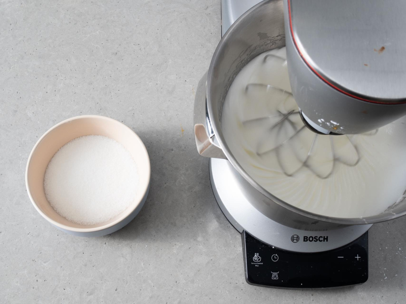 Ubijające się białka w misie miksera kuchennego. Obok w miseczce cukier.