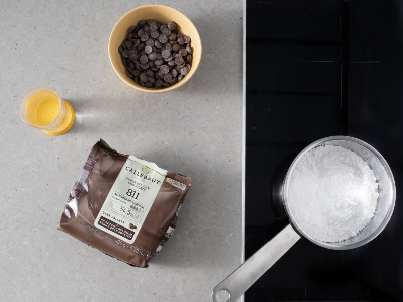 Na blacie kuchennym składniki potrzebne do przygotowania polewy: czekolada, woda i cukier puder.