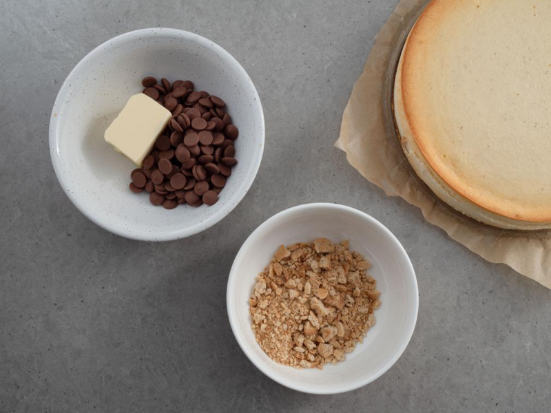 W miseczkach pokruszone ciasteczka obok kaletki czekolady Callebaut  z mas艂em i sernik na ciasteczkowym spodzie