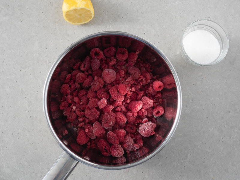 W garnku maliny. Obok połówka cytryny i cukier w miseczce.