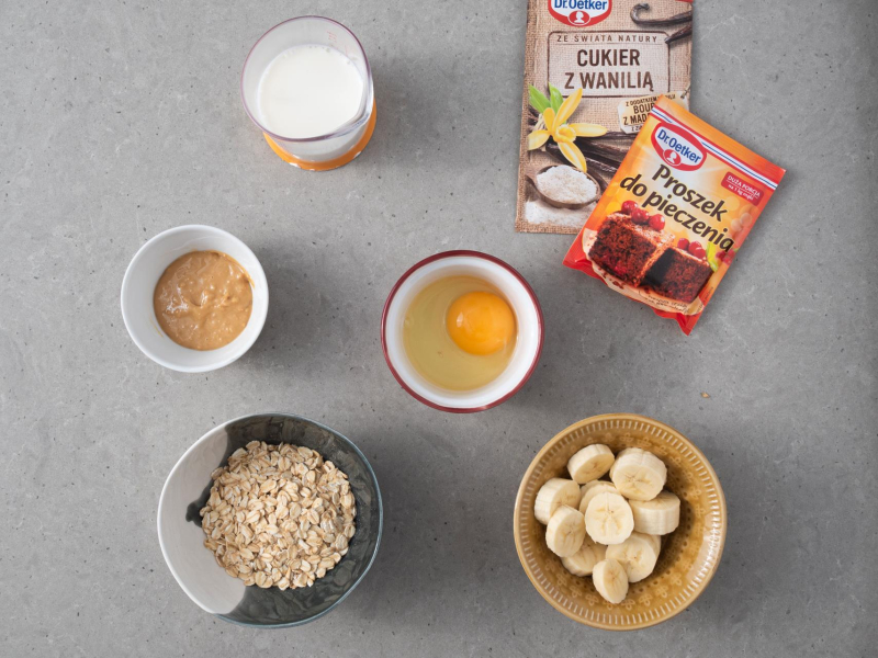 Na blacie miseczka z płatkami owsianymi, pokrojonymi bananami, jajkiem, proszkiem do pieczenia, cukrem  z wanilią, masłem orzechowym i mlekiem.