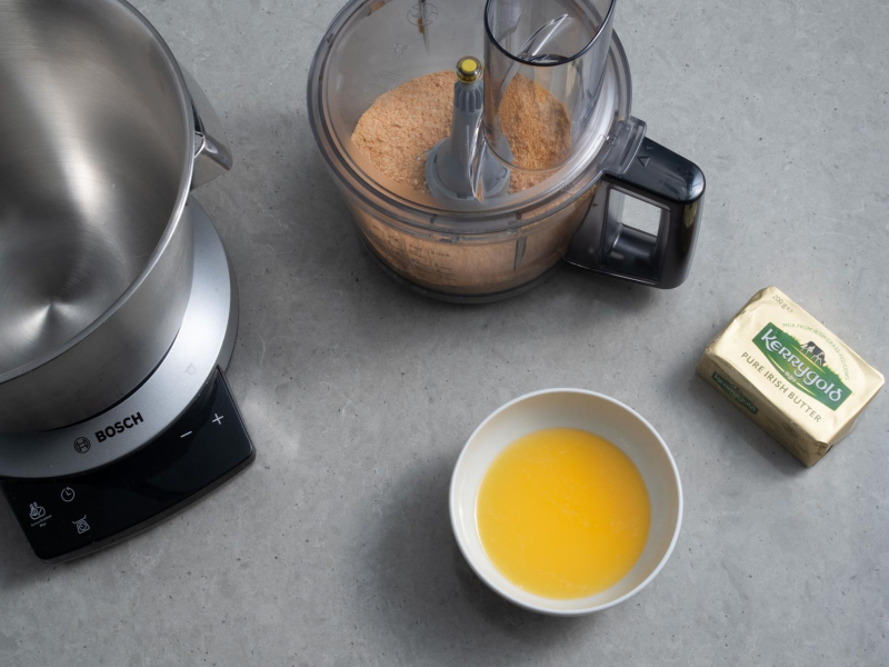 Blender ręczny firmy Bosch leżący na blacie kuchennym, w nim herbatniki. Obok miseczka z roztopionym masłem i kostka Kerrygold.