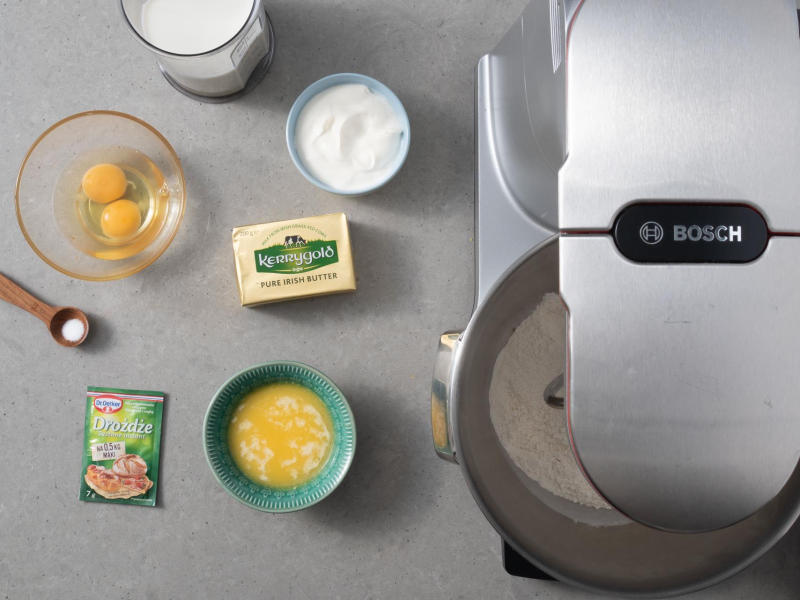 Na blacie kuchennym kostka masła, obok rozpuszczone masło w miseczce, 2 jajka w miseczce, kwaśna śmietana oraz mleko i suszone drożdże