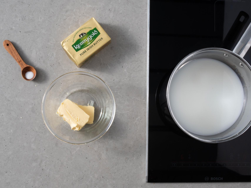 W garnku mleko z wodą. Obok kostka masła. Szczypta soli i cukru na łyżeczce i pokrojone masło z misce.