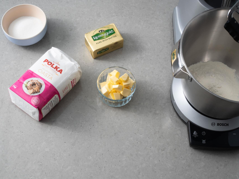 Na blacie kuchennym mąka, kostka masła oraz masło w miseczce. Obok robot kuchenny.