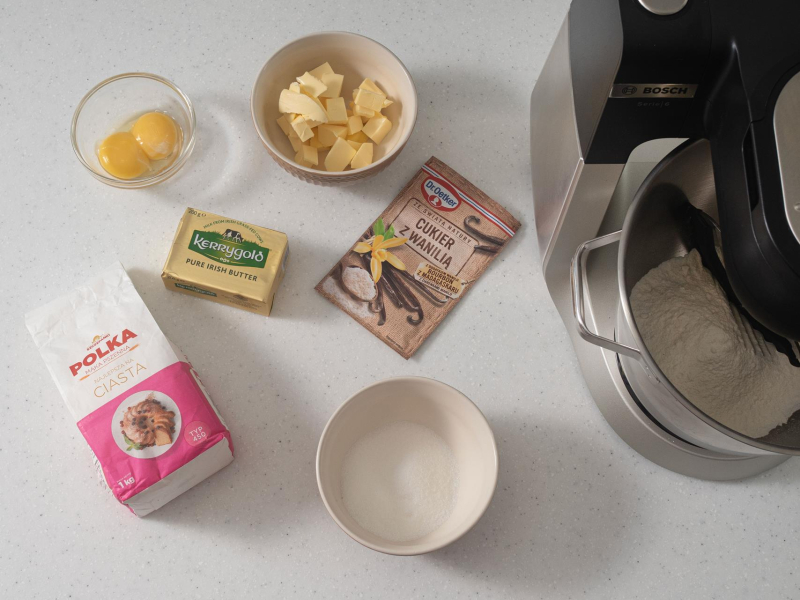 Na blacie kuchennym misa z mąką pszenną, cukier w misce, pokrojone masło oraz żółtka.