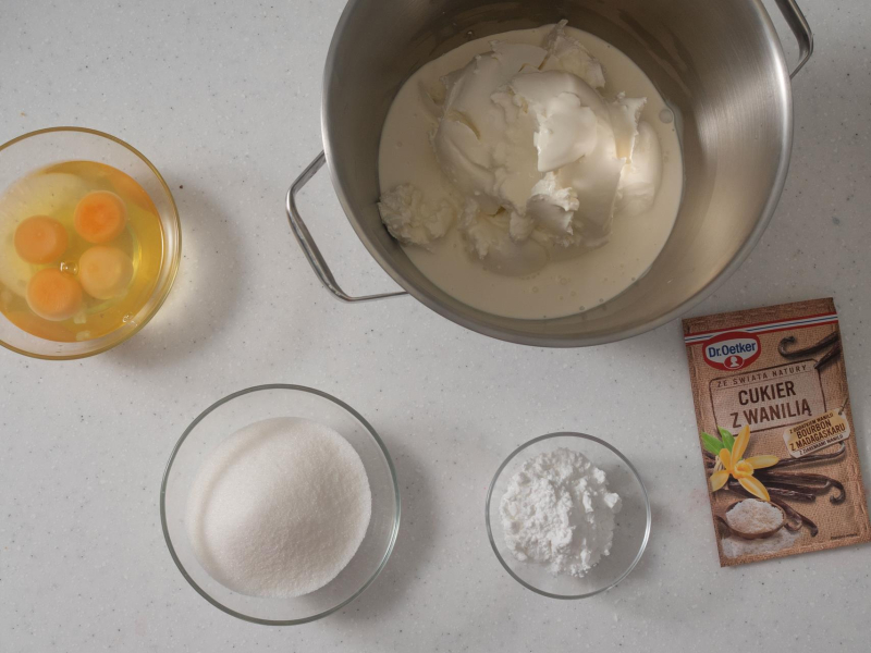 Twaróg sernikowy w dużej misie. Obok cukier, mąka ziemniaczana, jajka oraz opakowanie cukru z wanilią.