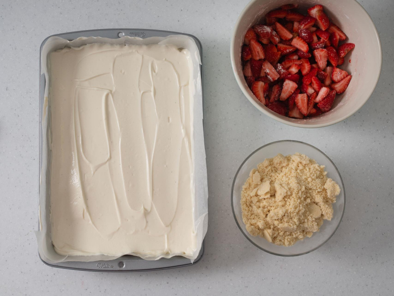 Masa serowa wyłożona w formie. Obok w misce pokrojone truskawki oraz kruszonka.