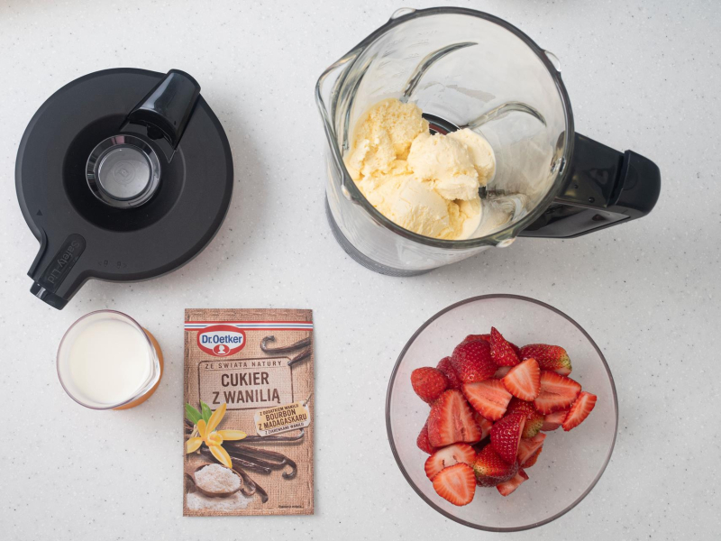 Na blacie kuchennym opakowanie cukru z wanilią, truskawki w miseczce, mleko oraz lody.