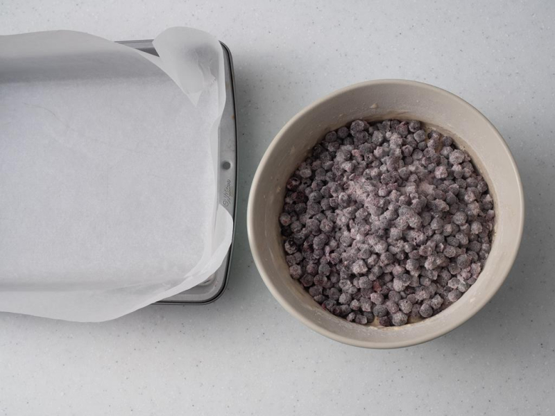 foremka do pieczenia wyłożona papierem do pieczenia obok miska z przygotowanym ciastem z jagodami.