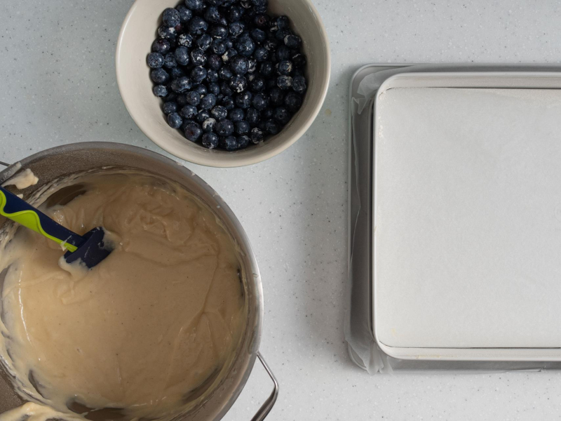 Forma wyłożona papierem do pieczenia. Obok w miseczce borówki i ciasto jogurtowe w misie.