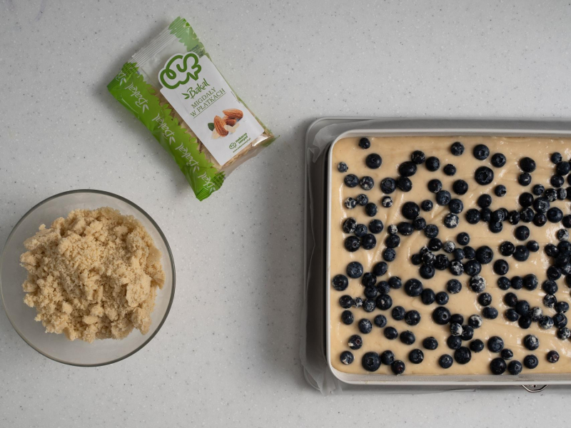 Ciasto przełożone do formy i posypane borówkami. Obok kruszonka i płatki migdałów w opakowaniu.