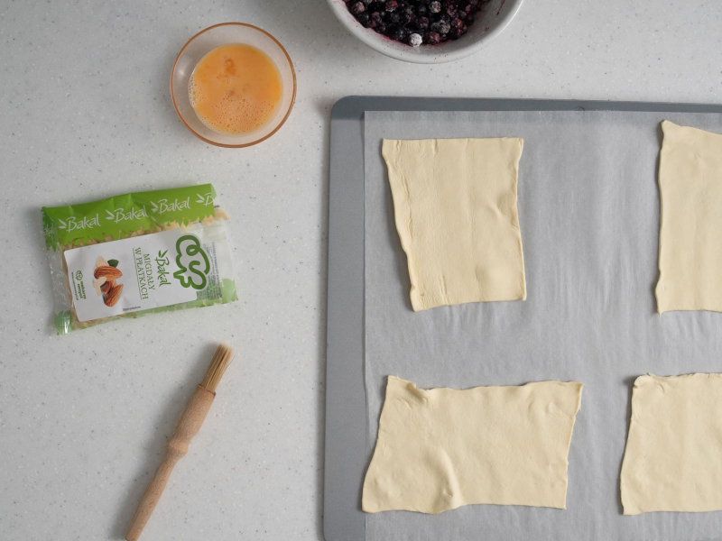 Na blasze rozłożony płaty ciasta francuskiego. Obok opakowanie migdałów w płatkach i roztrzepane jajko w miseczce.