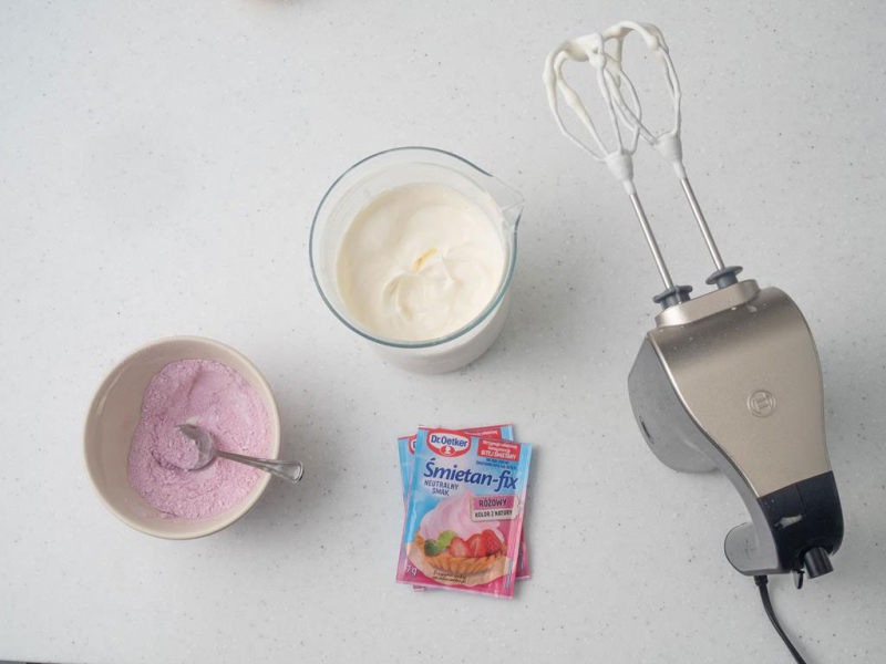 w szklanym naczyniu ubita śmietanka mikserem ręcznym obok różowe śmietan fixy