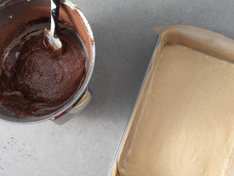 Jasne ciasto przełożone do formy. Obok ciasto czekoladowe w misie.