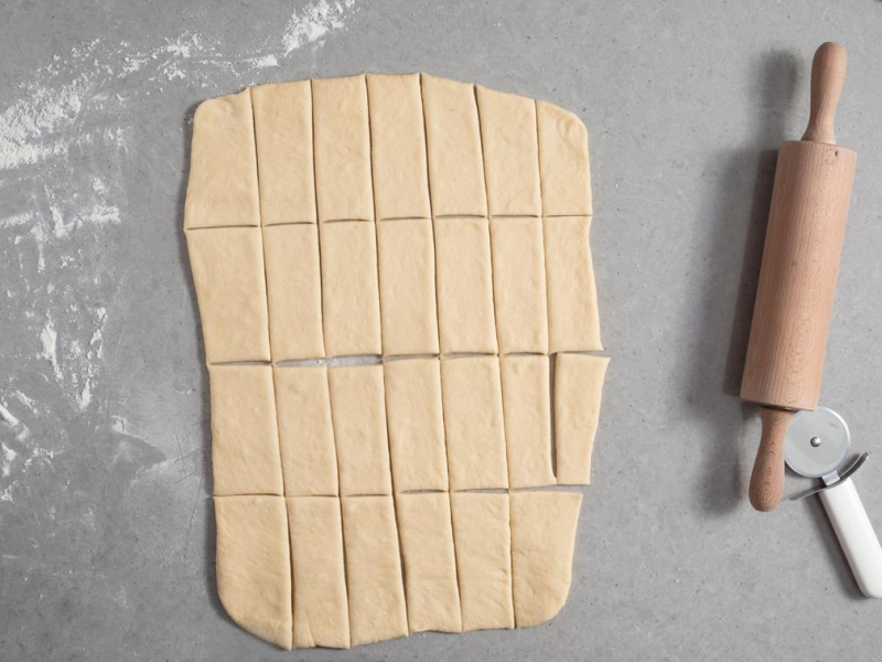 rozwałkowane ciasto drożdżowe pokrojone na prostokąty obok wałek i nóż do pizzy