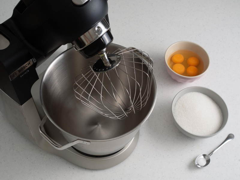 Robot kuchenny na blacie. Obok w miseczce jajka, cukier oraz sól na łyżeczce.