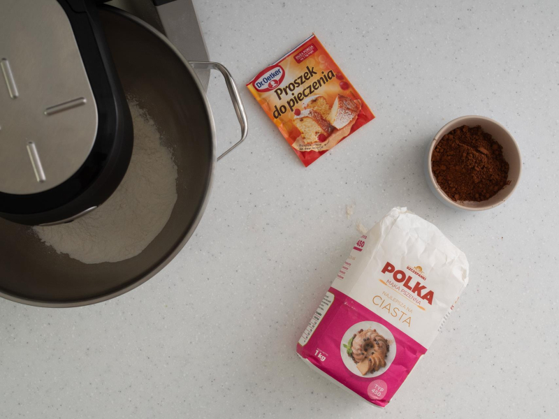 Na blacie kuchennym opakowanie mąki pszennej, proszek do pieczenia i kakao w miseczce.