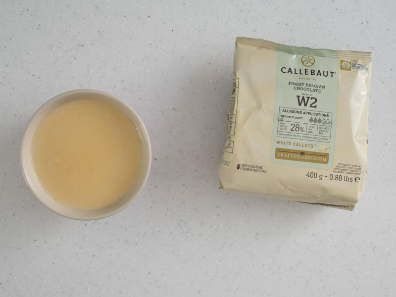Na blacie kuchennym opakowanie białej czekolady Callebaut W2, a obok w miseczce rozpuszczona czekolada biała ze śmietanką.