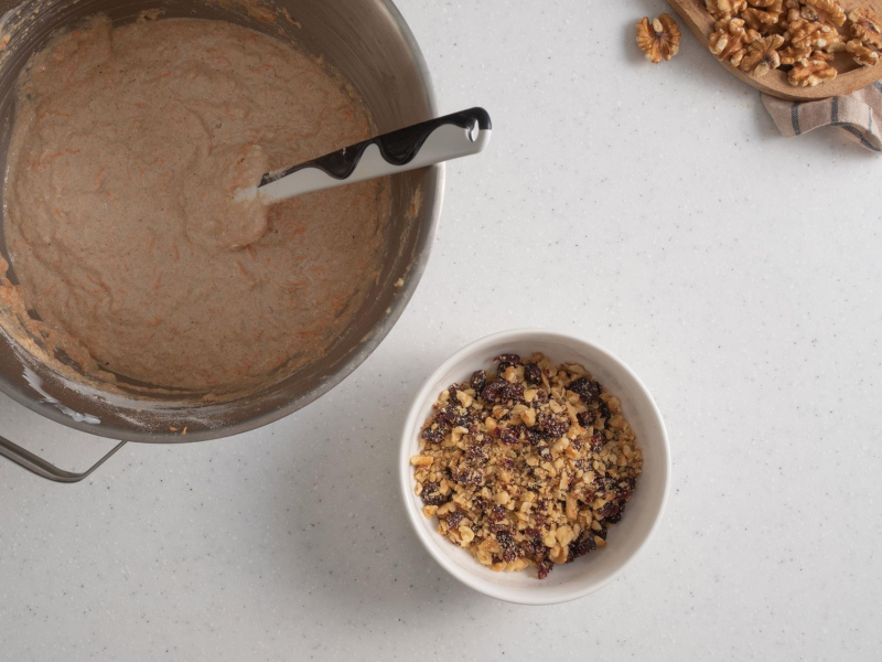 Surowe ciasto marchewkowe w dużej misie, a w małej posiekane orzechy i żurawina.