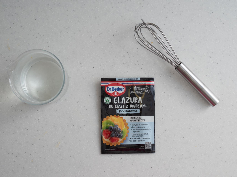 Na blacie kuchennym opakowanie od glazury, wrzątek w szklanym naczyniu oraz trzepaczka.