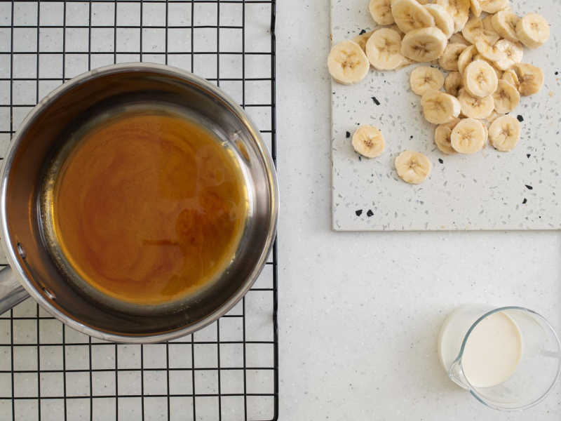 W garnku karmel. Obok pokrojone banany oraz śmietanka.