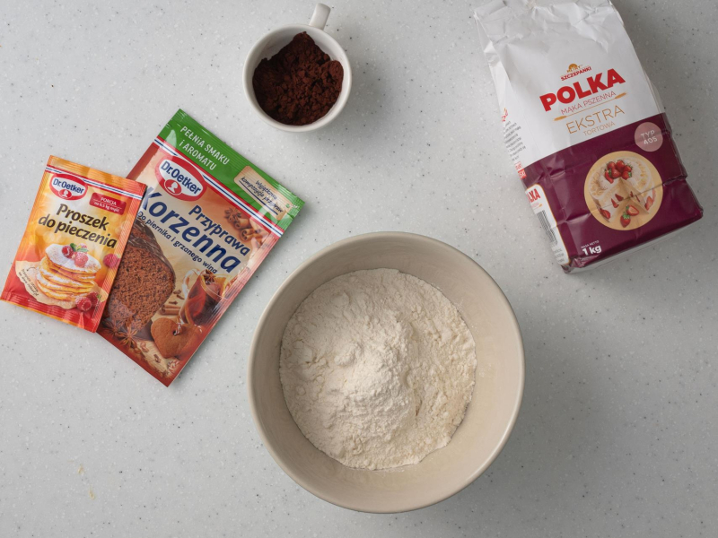 Na blacie kuchennym suche składniki do przygotowania ciasta: mąka, kakao, przyprawa korzenna oraz proszek do pieczenia.