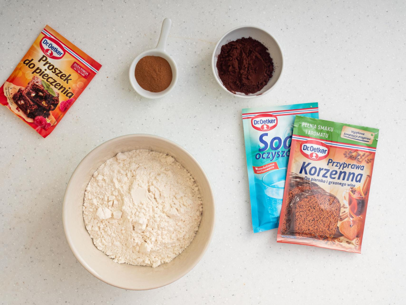 W misce mąka pszenna. Obok w miseczce kakao oraz cynamon. Na blacie opakowanie sody, przyprawy korzennej oraz proszku do pieczenia.