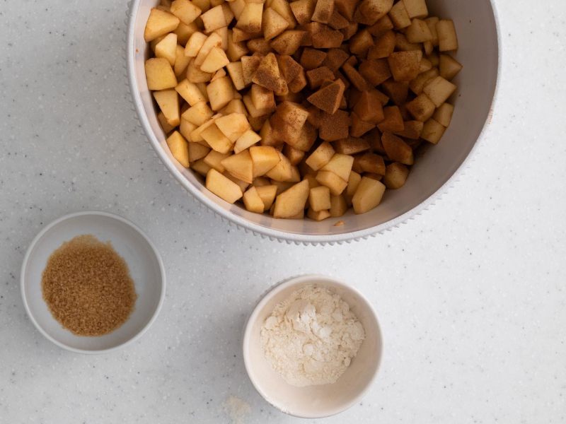 Pokrojone jabłka z cynamonem w misce. Obok cukier trzcinowy i mąka w miseczkach.