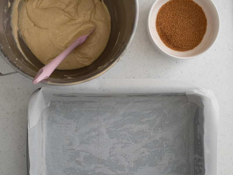 Forma wyłożona papierem do pieczenia. Obok surowe ciasto i posypka cynamonowa.