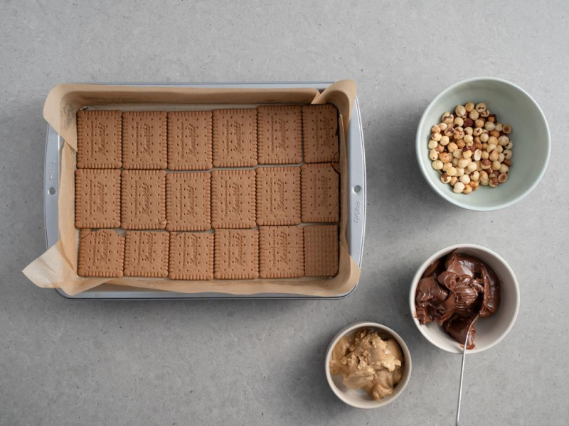 forma wyłożona herbatnikami, w miseczce nutella, masło orzechowe i orzechy laskowe