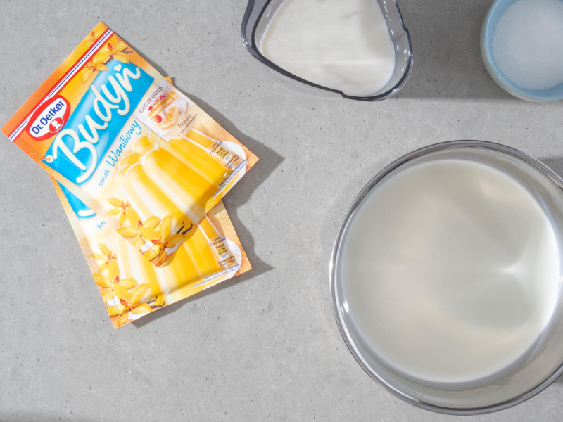 W garnku mleko. Obok opakowania budyniu o smaku waniliowym Dr. Oetkera.