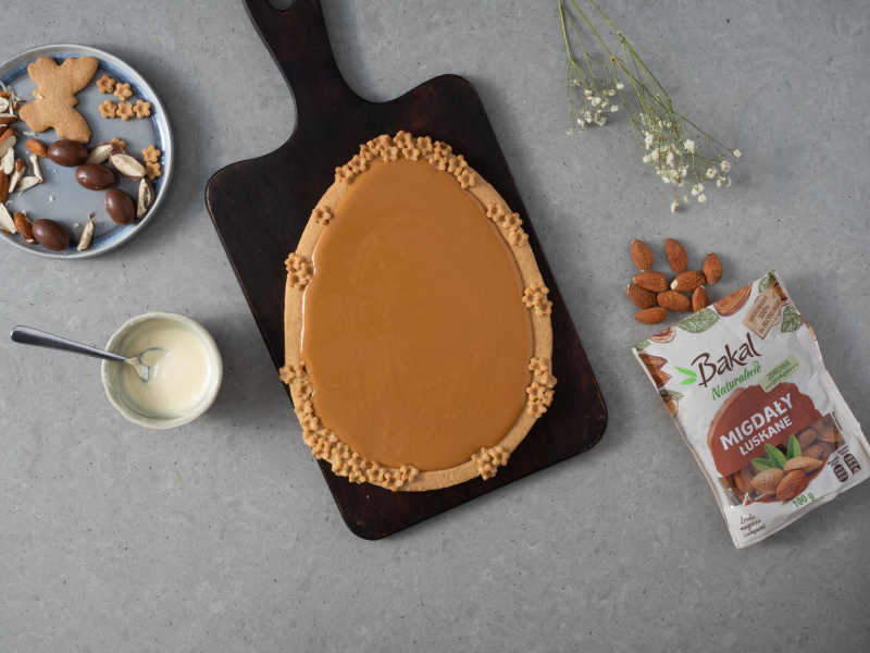 Mazurek bezglutenowy z masą kajmakową na drewnianej desce. Obok migdały Bakal, rozpuszczona biała czekoladowa oraz dodatki do dekoracji.